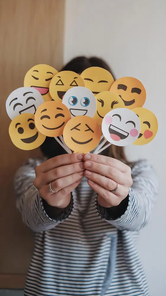 Osoba trzymająca przed twarzą papierowe wycinanki różnych emotikonów, ubrana w rękawy w paski.