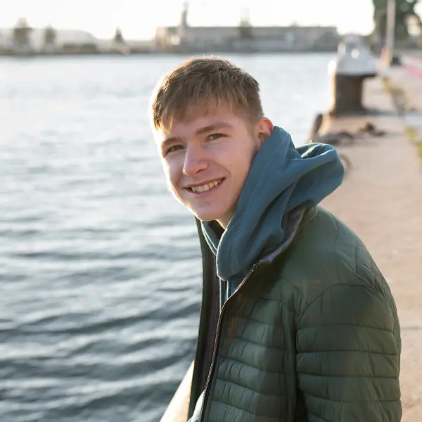 Uśmiechnięty młody mężczyzna w kurtce nad wodą w słoneczny dzień.