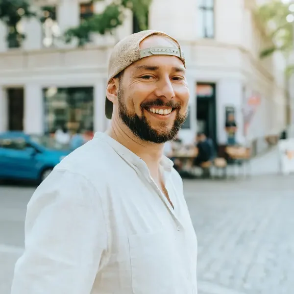 Uśmiechnięty mężczyzna w jasnej koszuli i czapce z daszkiem do tyłu stoi na miejskiej ulicy, w tle widać ogródek restauracyjny.