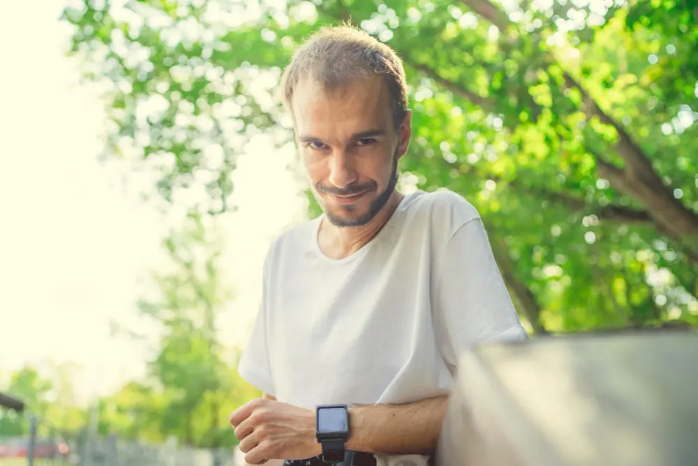 Uśmiechnięty mężczyzna w białej koszuli stoi na zewnątrz, przy drewnianej barierce, a w tle widać zielone drzewa.