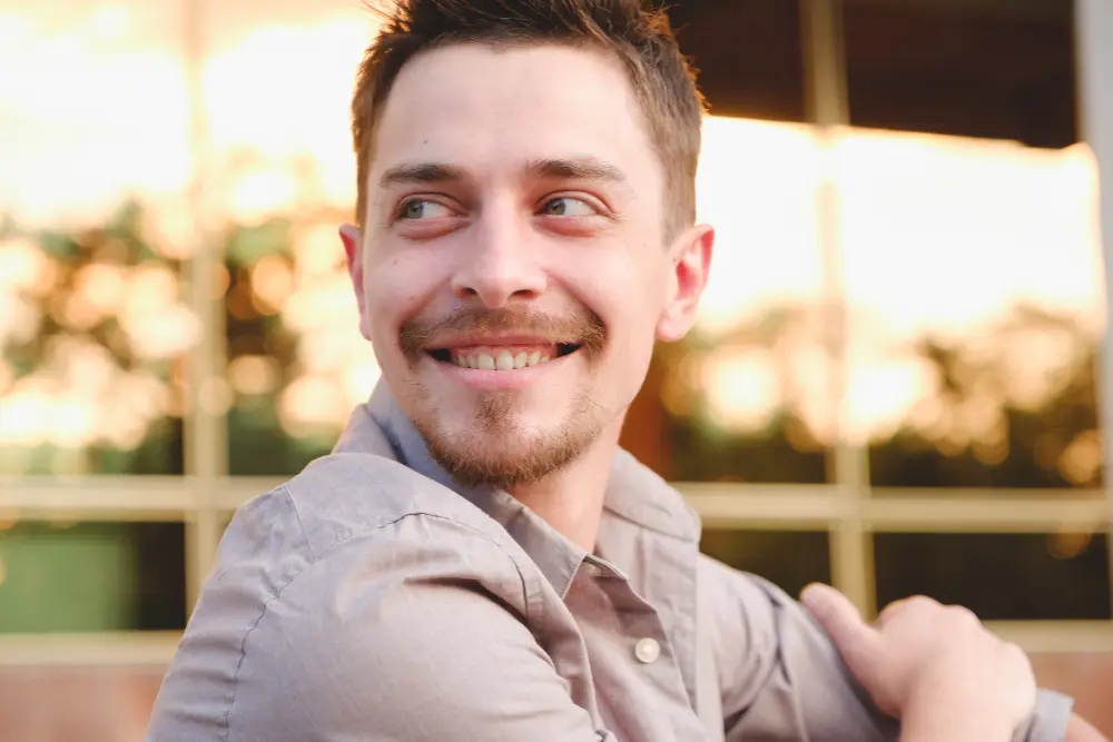 Uśmiechnięty mężczyzna z krótkimi włosami i bródką, patrzący w lewo, na zewnątrz, w tle widać słońce.