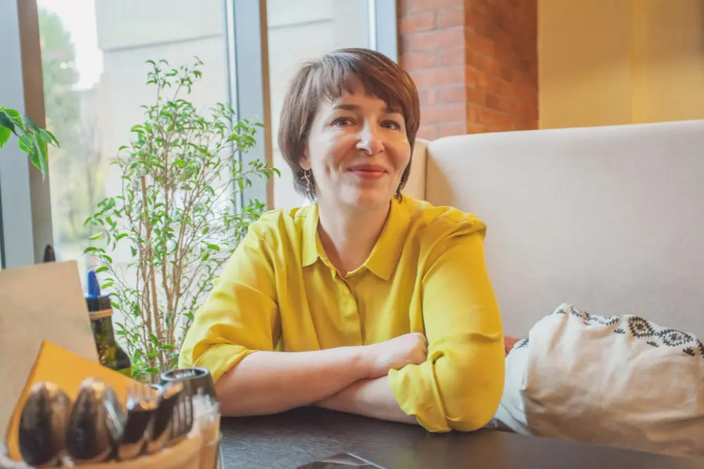 Kobieta w żółtej koszulce uśmiecha się, siedząc przy stoliku w przytulnej, słonecznej kawiarni.