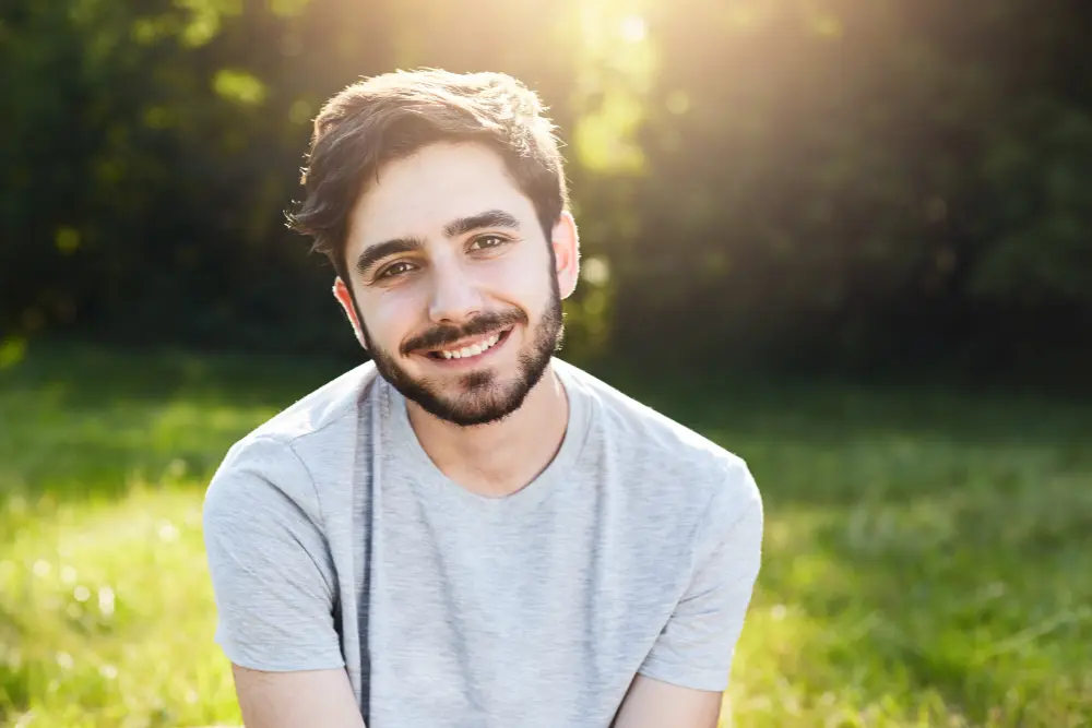 Młody mężczyzna z brodą uśmiecha się, siedząc na trawie w słońcu, w tle widać drzewa.