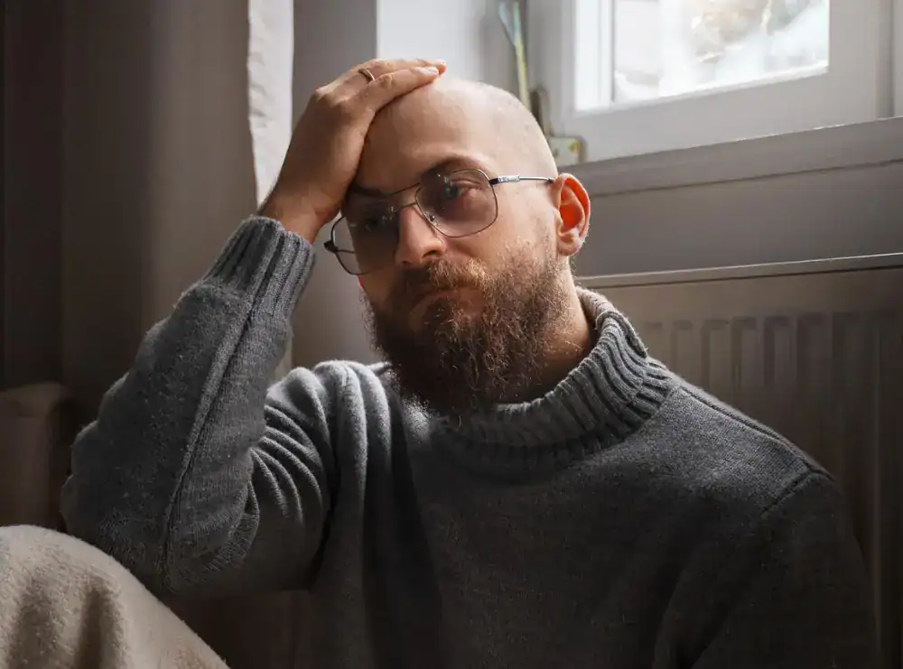 Brodaty mężczyzna w okularach i szarym swetrze siedzi przy oknie, opierając głowę na ręce i wygląda na zamyślonego.