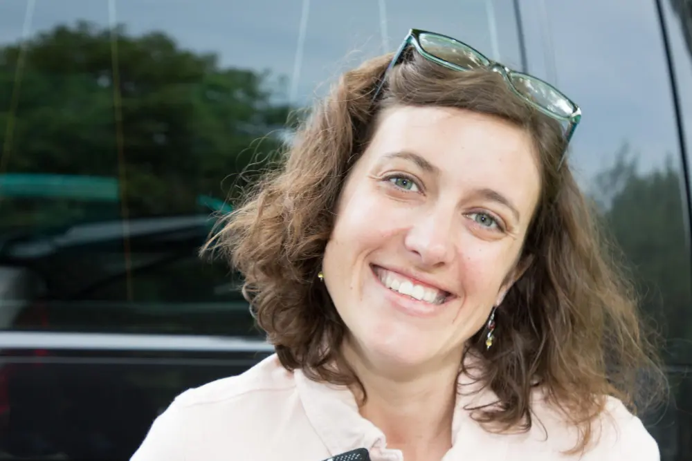 Uśmiechnięta kobieta z falowanymi brązowymi włosami, w okularach na głowie, stojąca przed samochodem.