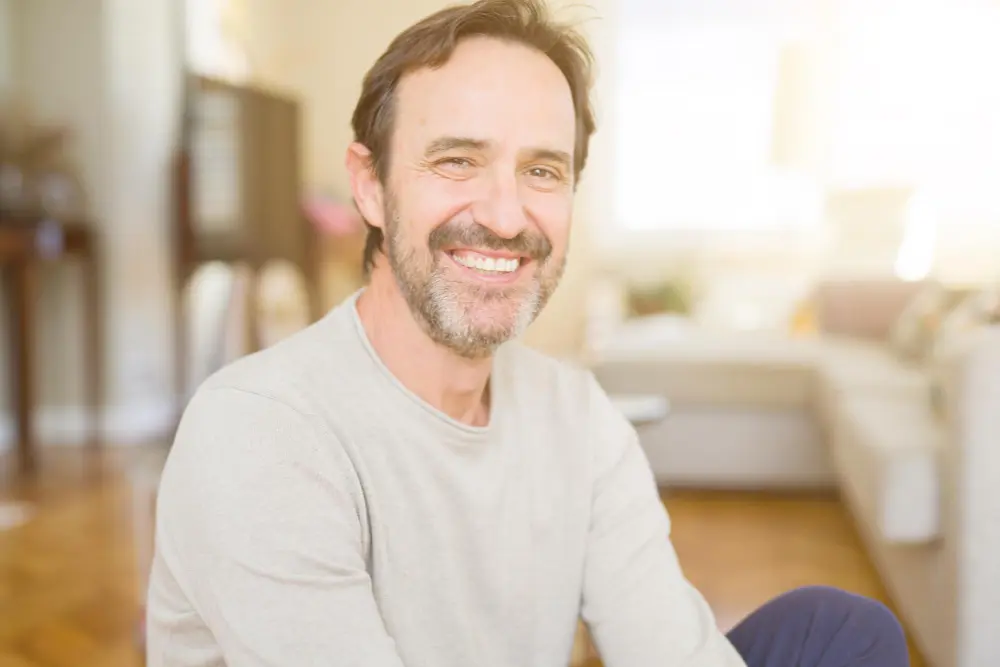 Uśmiechnięty mężczyzna o brązowych włosach i brodzie siedzi w jasnym salonie z rozmytym tłem.