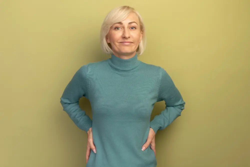 Uśmiechnięta kobieta z krótkimi blond włosami w turkusowym swetrze stoi z rękami na biodrach na zielonym tle.