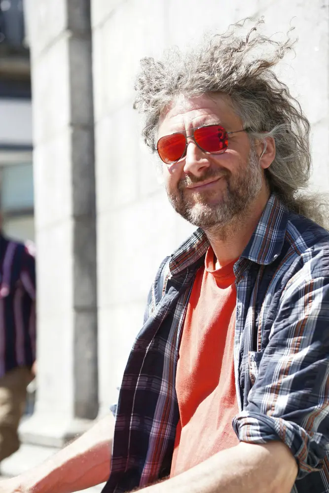 Mężczyzna z kręconymi siwymi włosami, czerwonymi okularami przeciwsłonecznymi, kraciastą koszulą i pomarańczowym T-shirtem uśmiecha się na zewnątrz w świetle słonecznym.
