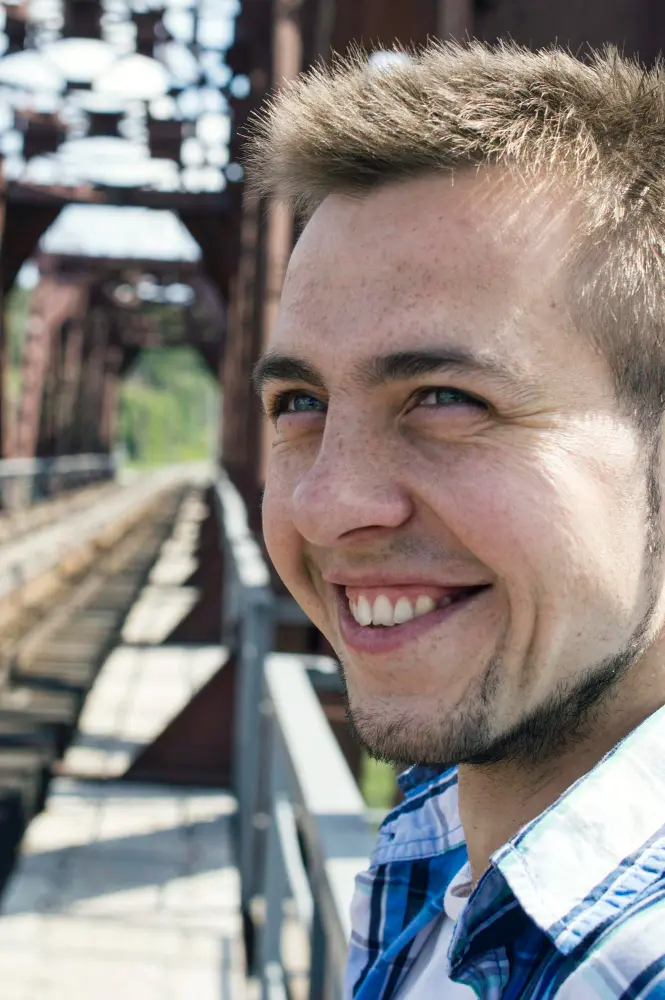 Uśmiechnięty mężczyzna z krótkimi włosami i zarostem stojący na moście, na tle metalowych belek i torów.