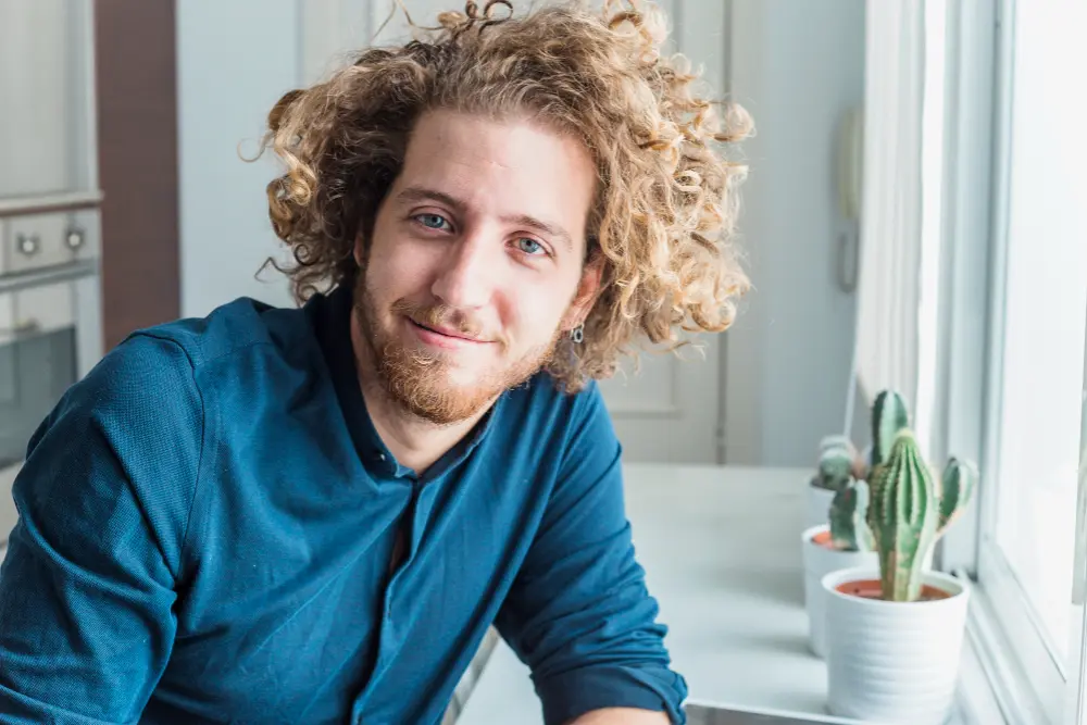 Mężczyzna z kręconymi włosami i brodą, uśmiechnięty, siedzi w domu przy oknie, na parapecie którego stoją doniczkowe kaktusy.