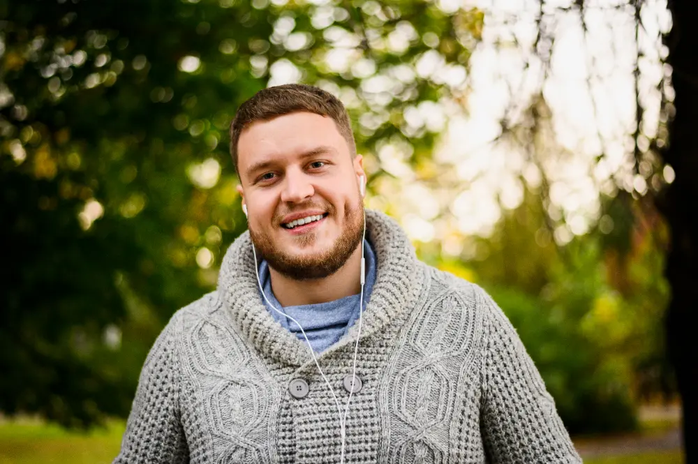 Uśmiechnięty mężczyzna w szarym swetrze ze słuchawkami, stojący na zewnątrz, w tle widać drzewa.