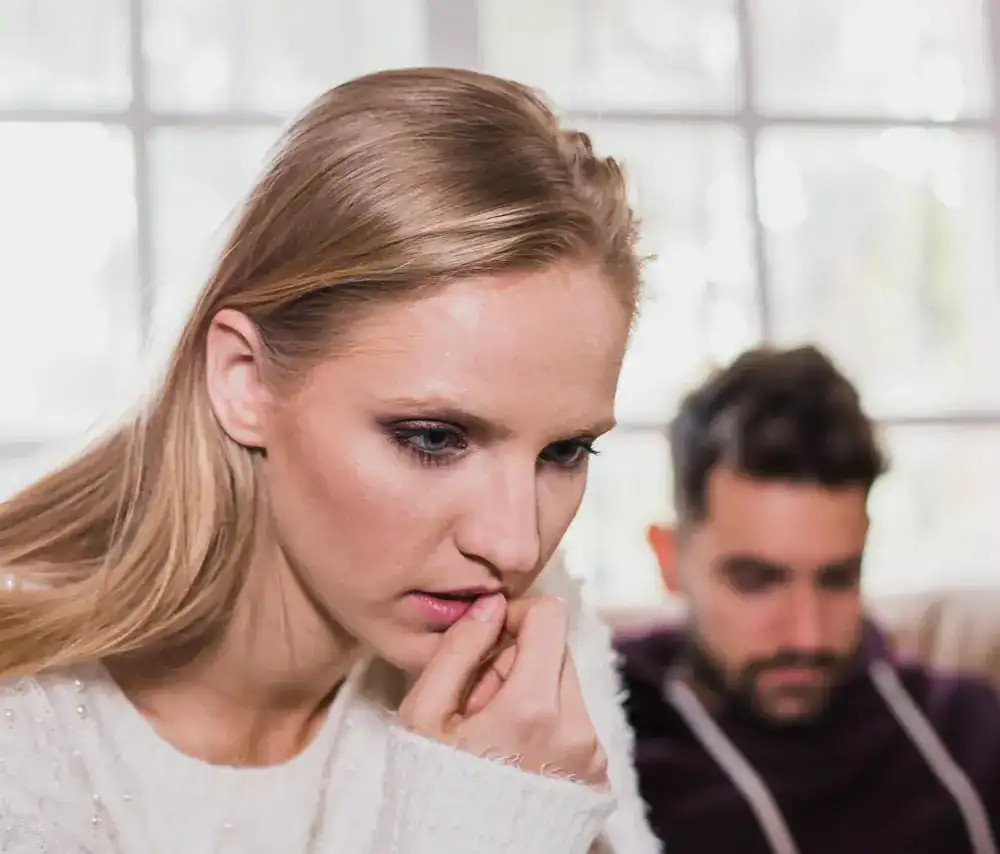 Zaniepokojona kobieta siedzi na pierwszym planie, podczas gdy mężczyzna siedzi w rozmazanym tle.