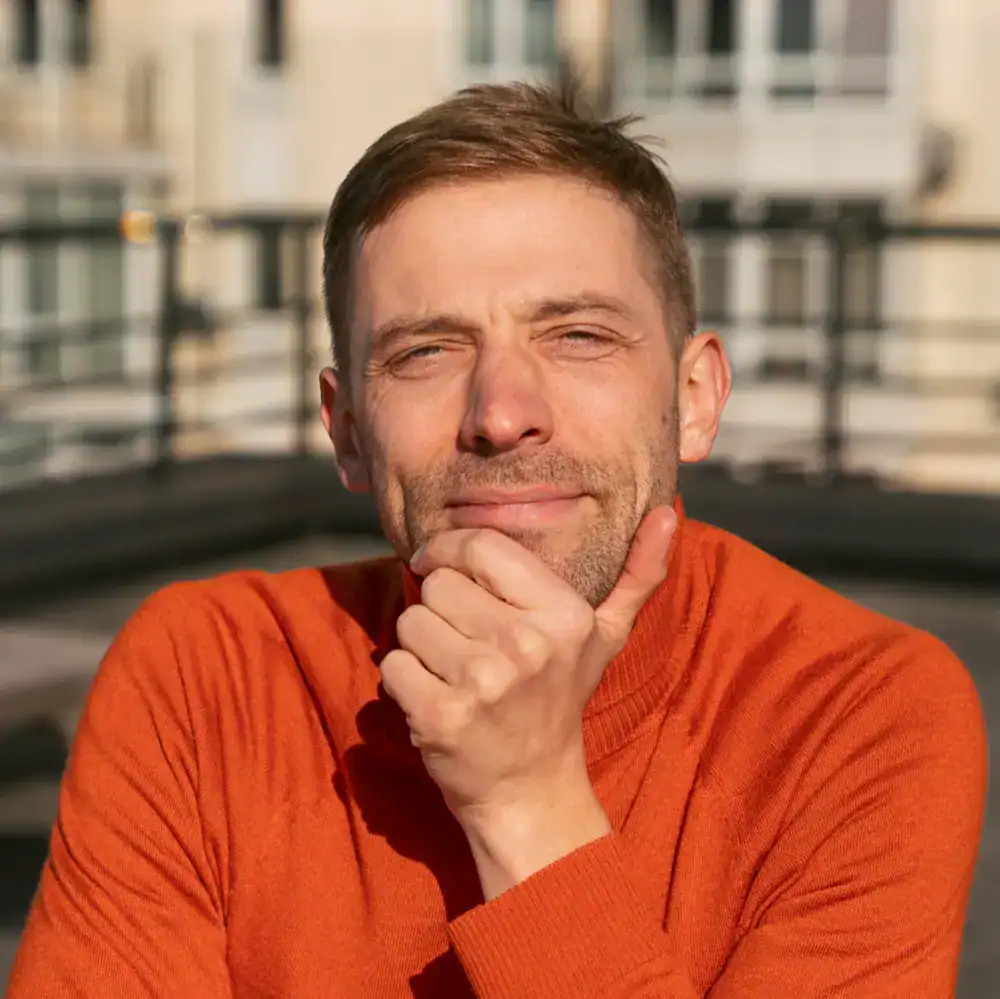 Mężczyzna w pomarańczowym swetrze siedzi na zewnątrz, opierając brodę na dłoni.