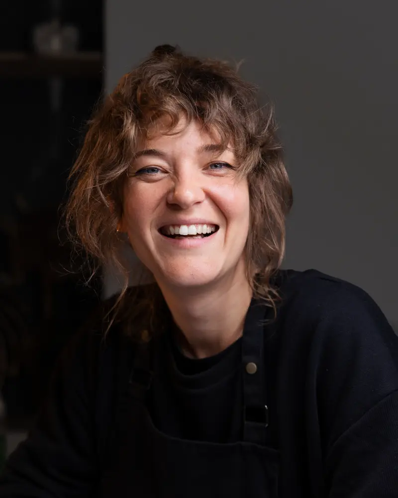 Uśmiechnięta kobieta z falowanymi włosami, ubrana w czarną koszulę i fartuch, siedzi w pomieszczeniu na ciemnym tle.