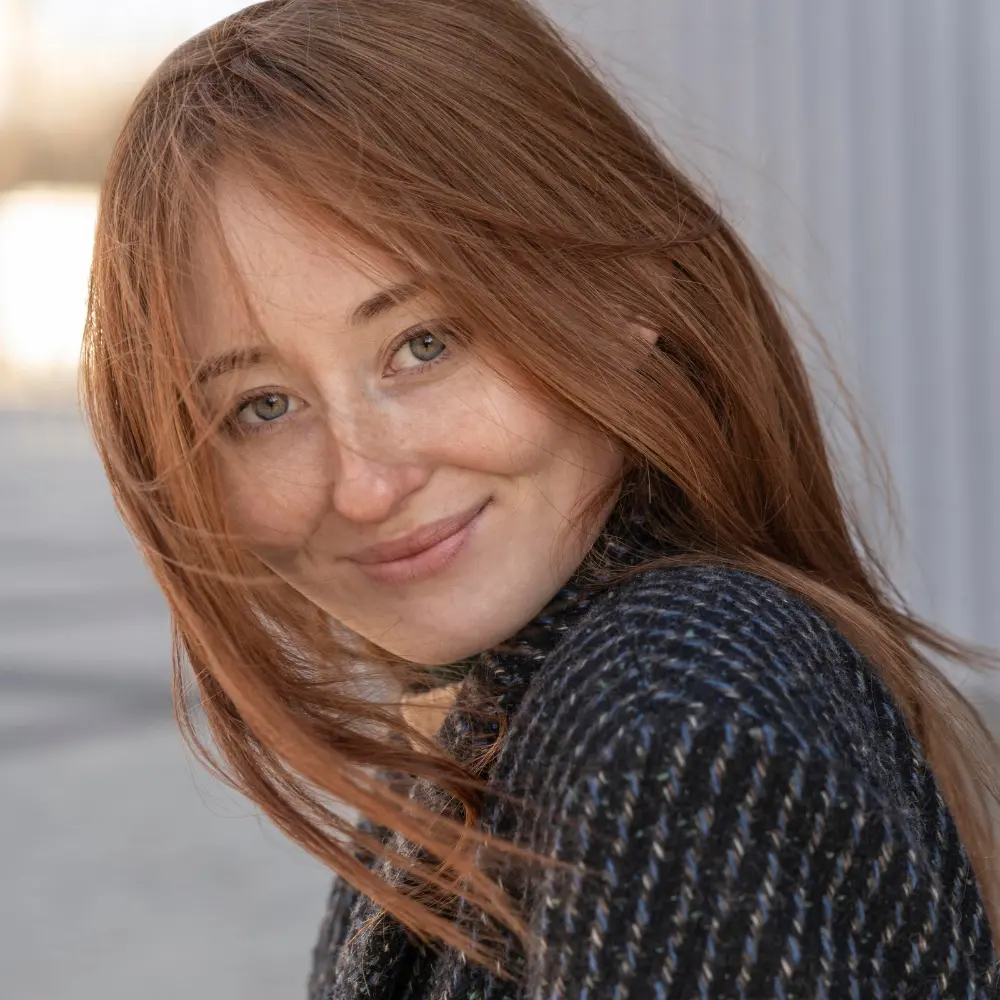 Kobieta z długimi, rudymi włosami uśmiecha się na zewnątrz, ubrana w ciemny, wzorzysty płaszcz.