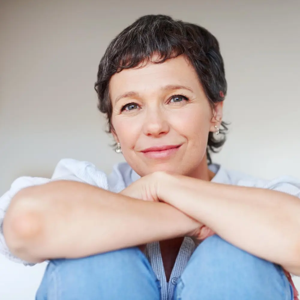 Uśmiechnięta kobieta o krótkich brązowych włosach siedzi z rękami opartymi na kolanach, wygląda na zrelaksowaną i zadowoloną.