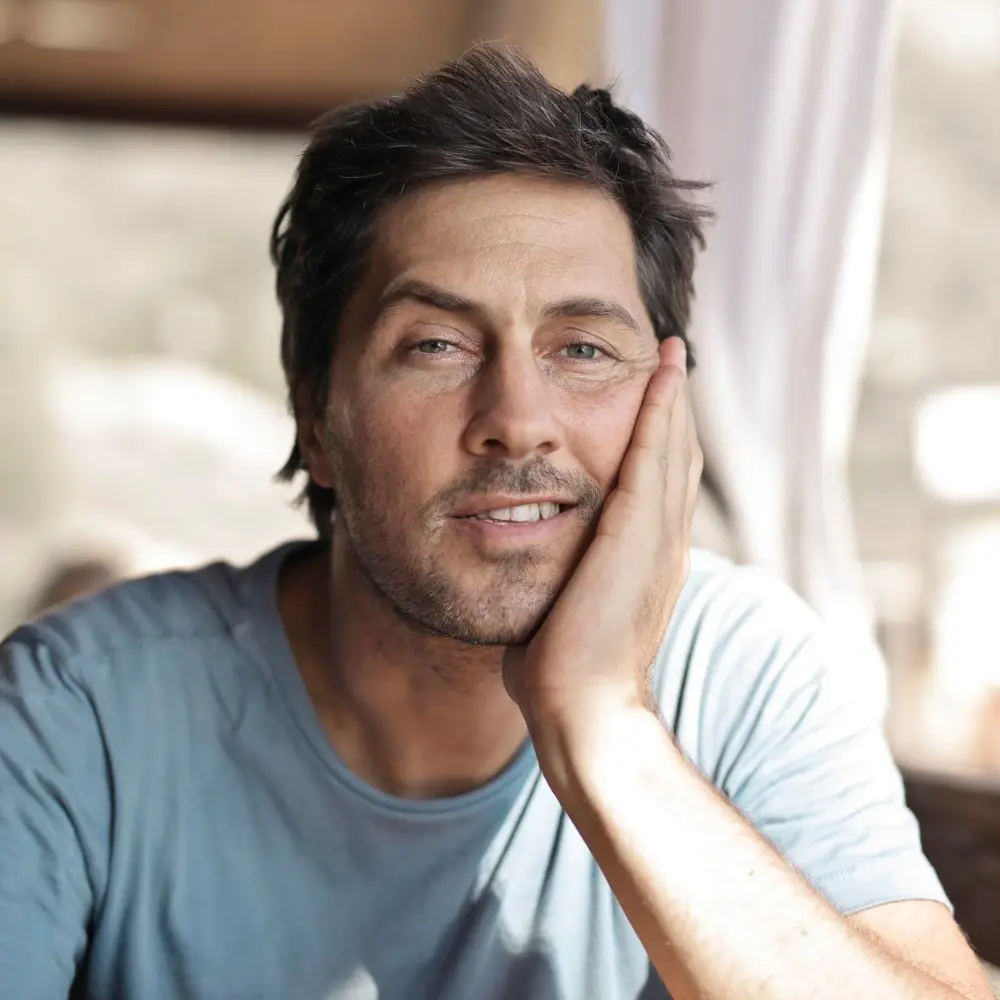 Mężczyzna o ciemnych włosach i niebieskiej koszuli opiera twarz na dłoni i patrzy w kamerę z lekkim uśmiechem.