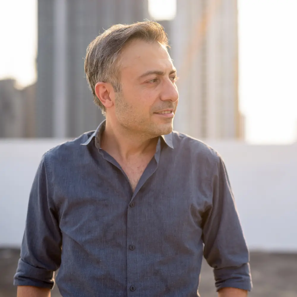 Mężczyzna w niebieskiej koszuli stoi na zewnątrz, patrzy w bok, w tle widać światło słoneczne i miejskie budynki.