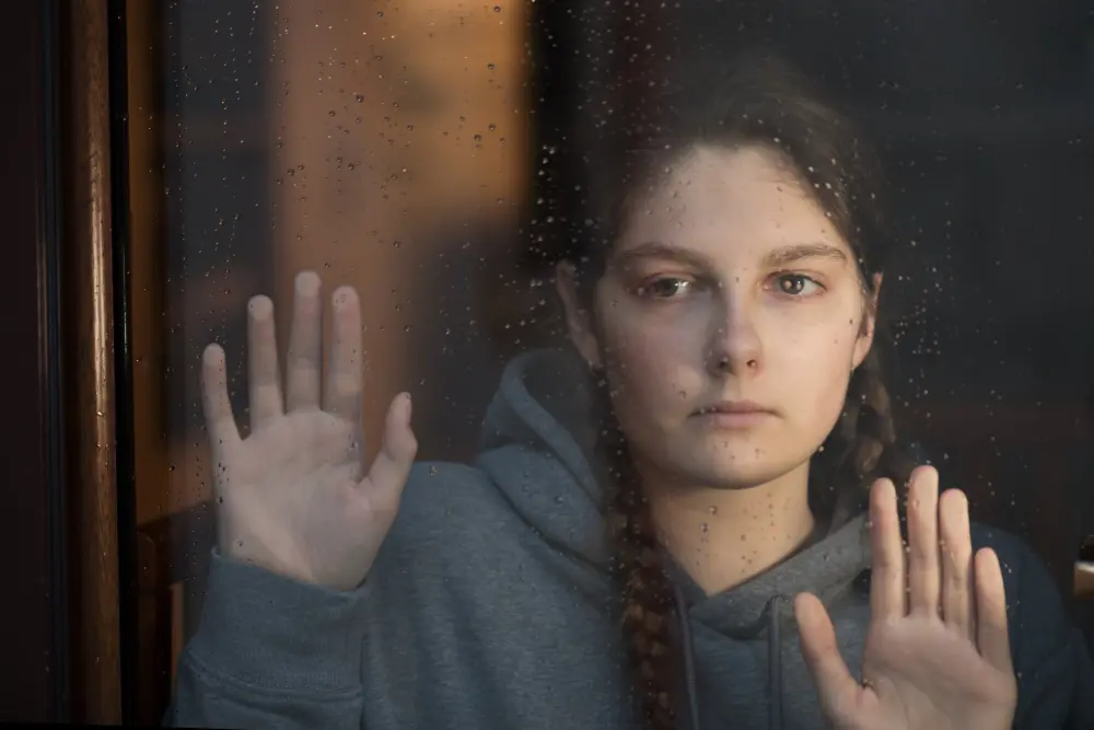 Młoda kobieta z warkoczami patrzy przez okno, przyciskając dłonie do szyby, w deszczowy dzień.