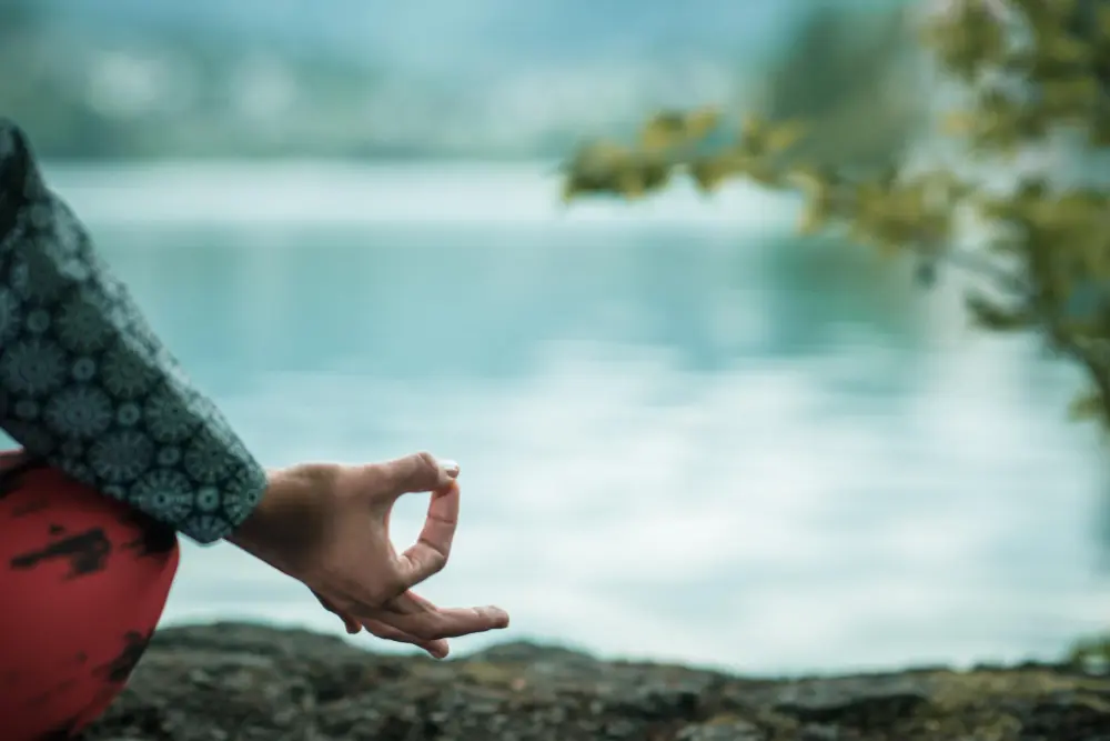 Ręka osoby w pozie medytacyjnej nad spokojnym jeziorem, z rozmytą wodą i zielenią w tle.